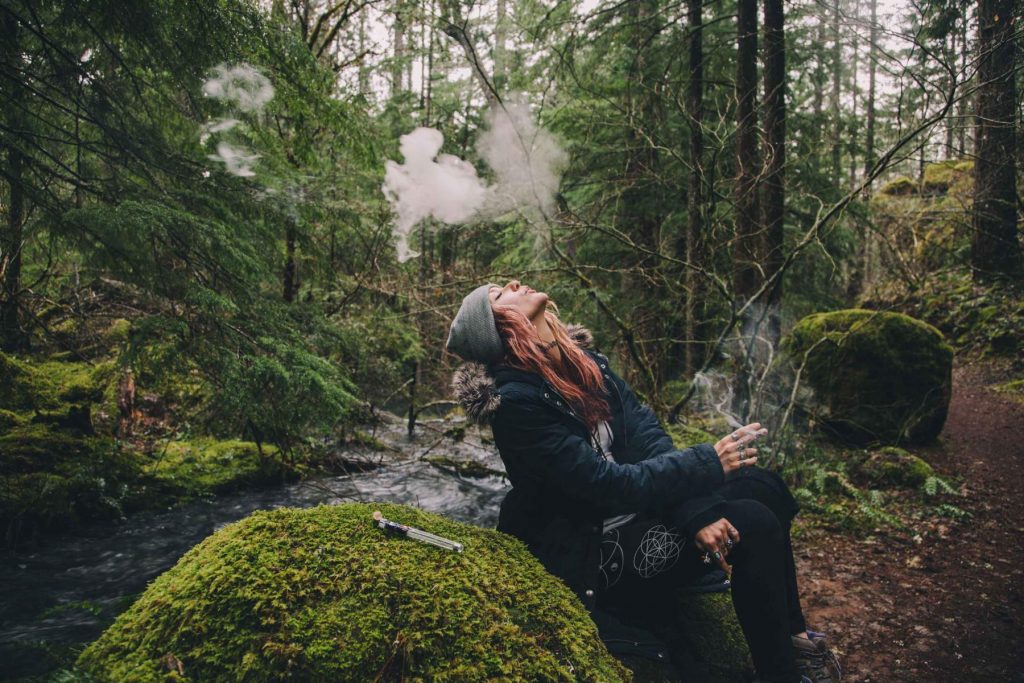 A person wearing a beanie and jacket exhales vapor while sitting on mossy ground in a forest near a stream. Smoke is visible, and a vaping device rests nearby.