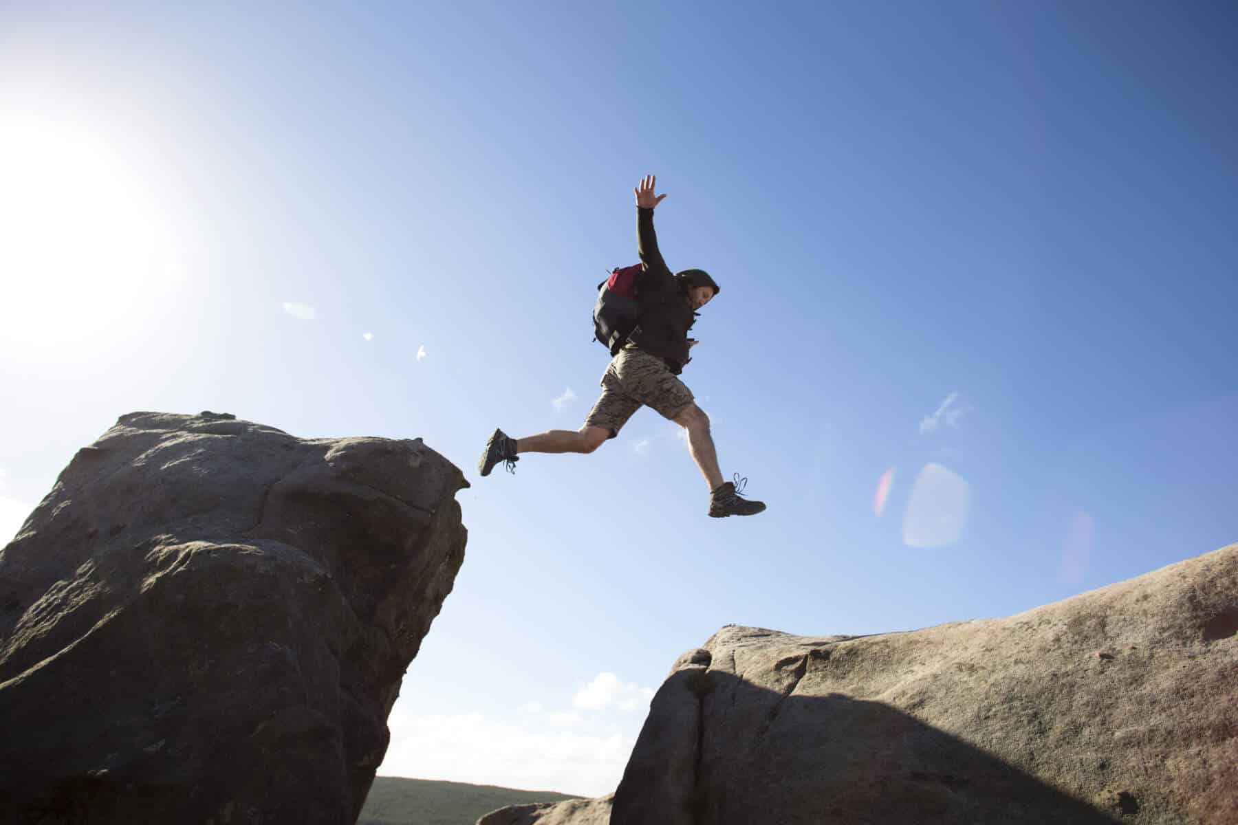 A person wearing a backpack and hat jumps between two large rocks under a clear blue sky.