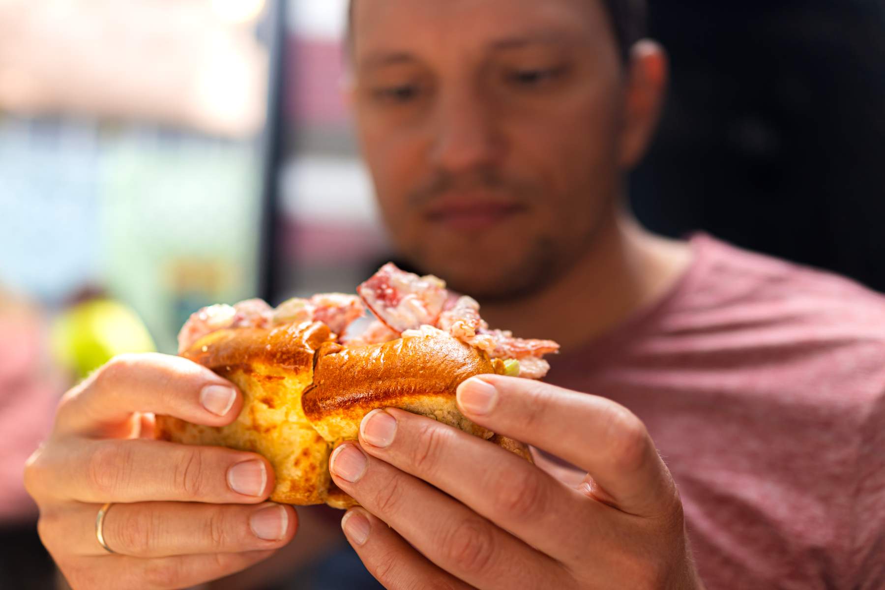 A man in a red shirt holds a lobster roll sandwich with both hands, his face slightly out of focus in the background.