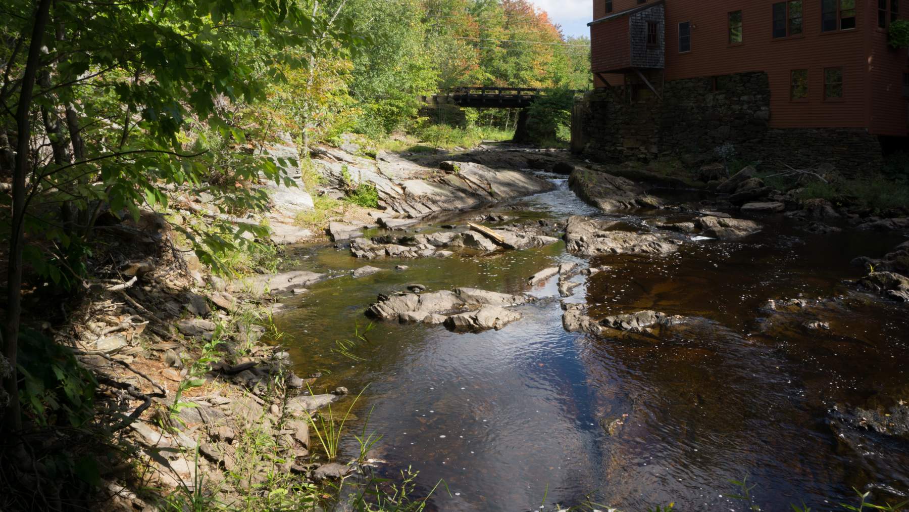 A small creek with clear water flows over rocks beside an old building and under a wooden bridge surrounded by lush greenery.