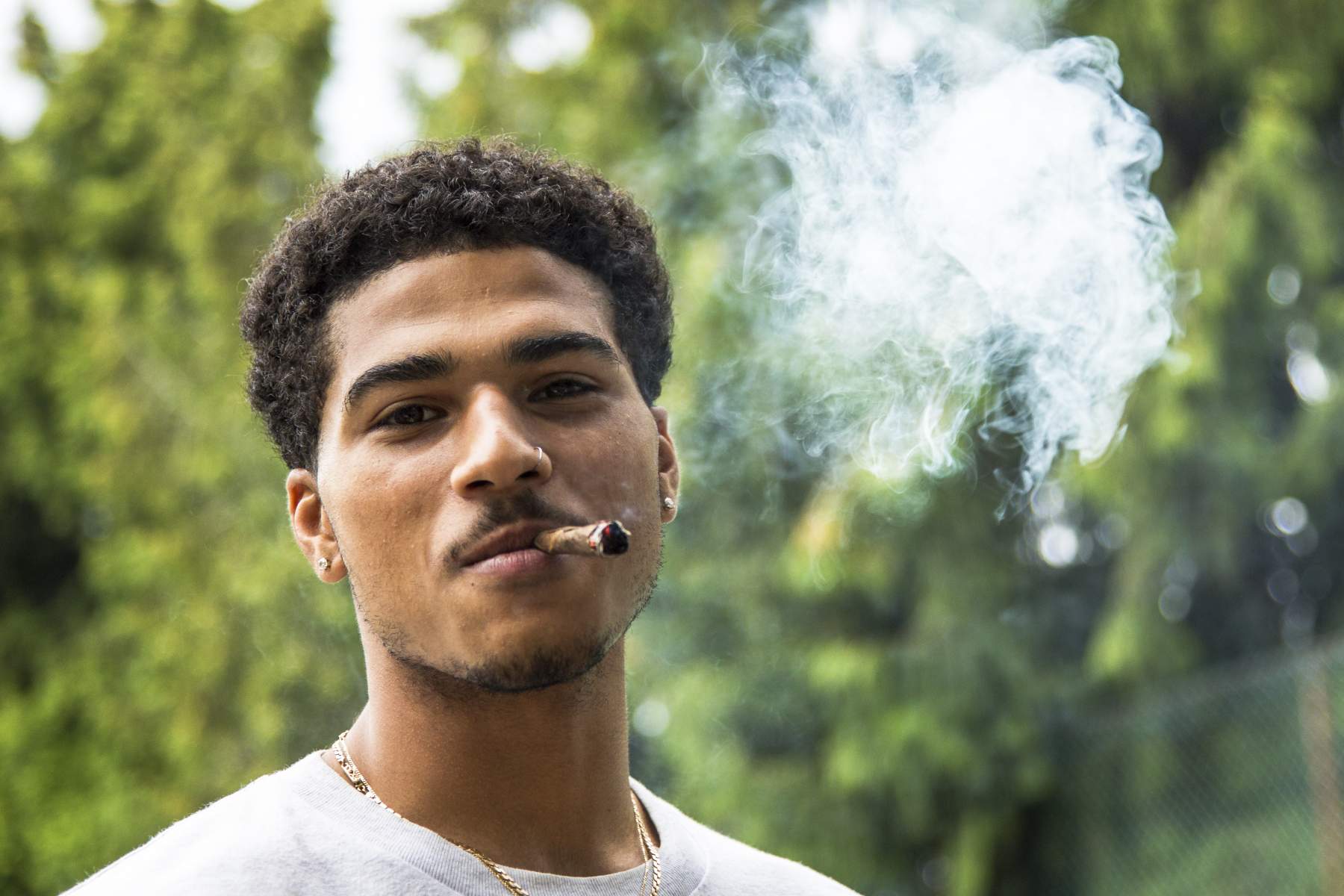 A man with short curly hair smokes a cigar outdoors, with smoke visible in the air. He wears a white t-shirt and a gold necklace. Trees are displayed in the blurred background.