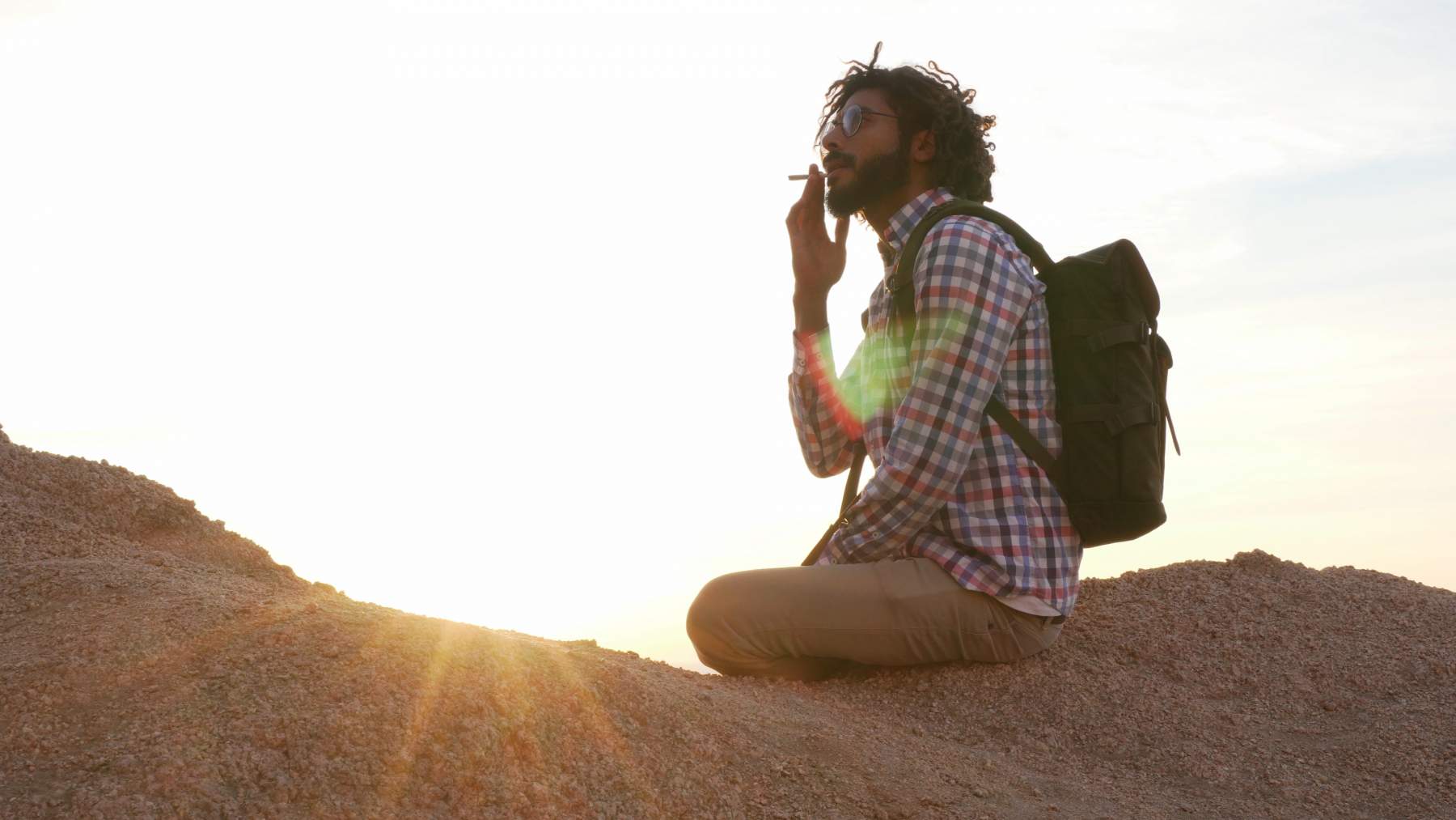 A person with a backpack and sunglasses kneels on a rocky hill at sunset, holding a small object near their mouth.