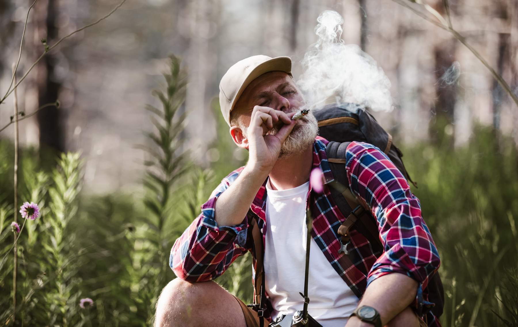 A person wearing a cap and plaid shirt is smoking while crouching in a forest. They have a backpack and a camera around their neck. Smoke is visible in the air.