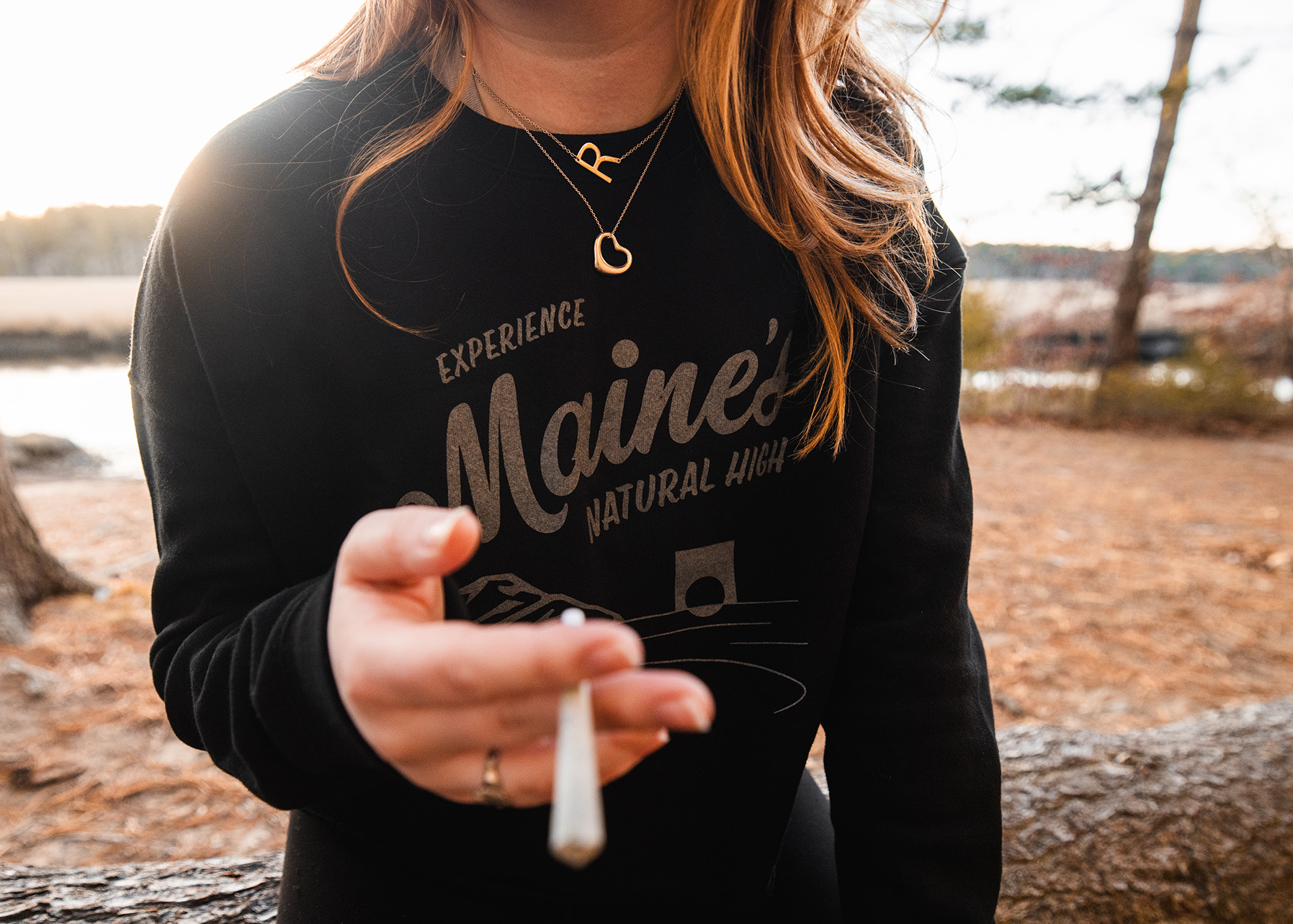 Woman holding a pre-roll joint and wearing a Maine's Natural High East Coast Cannabis crop top while outside in a Maine marsh.