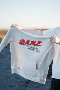 Person holding a beige hoodie that reads "DARE TO LIVE THE ADVENTURE" in red and black text.