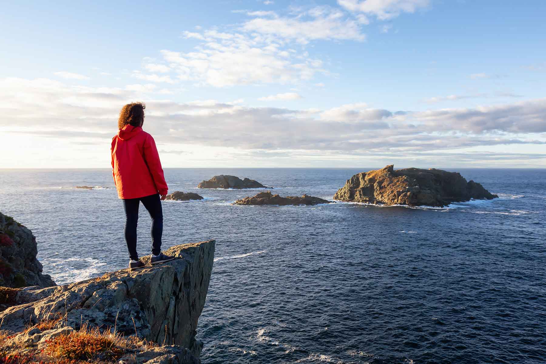 A person in a red jacket stands on a cliff overlooking the ocean with rocky islands under a partly cloudy sky.