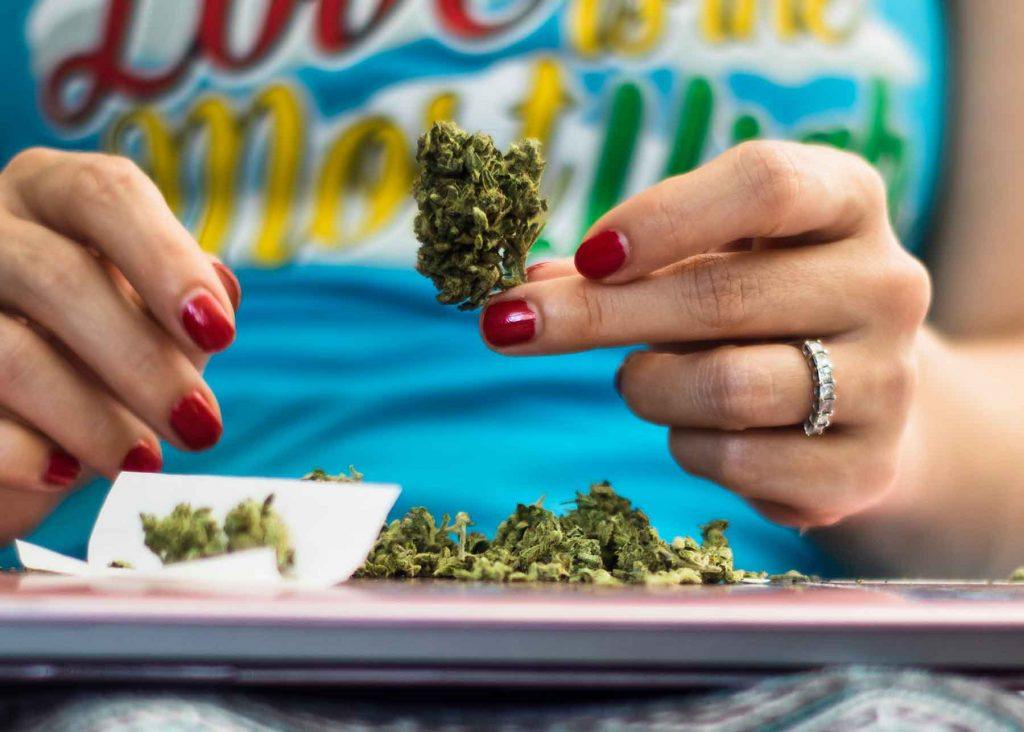 A person with red nail polish and a ring on their finger holding a cannabis bud, with more buds and rolling paper on a surface below.