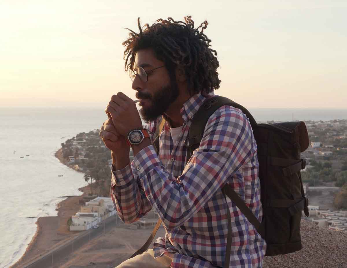 A man with glasses and a backpack, wearing a plaid shirt, is lighting a cigarette while sitting outdoors overlooking a coastal landscape at sunset.