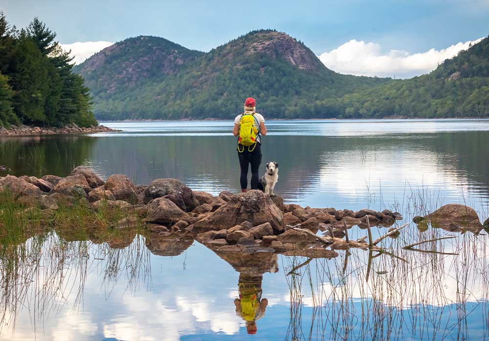 A person wearing a backpack stands with a dog on rocky ground by a calm lake, with mountains, trees, and a mostly cloudy sky in the background.